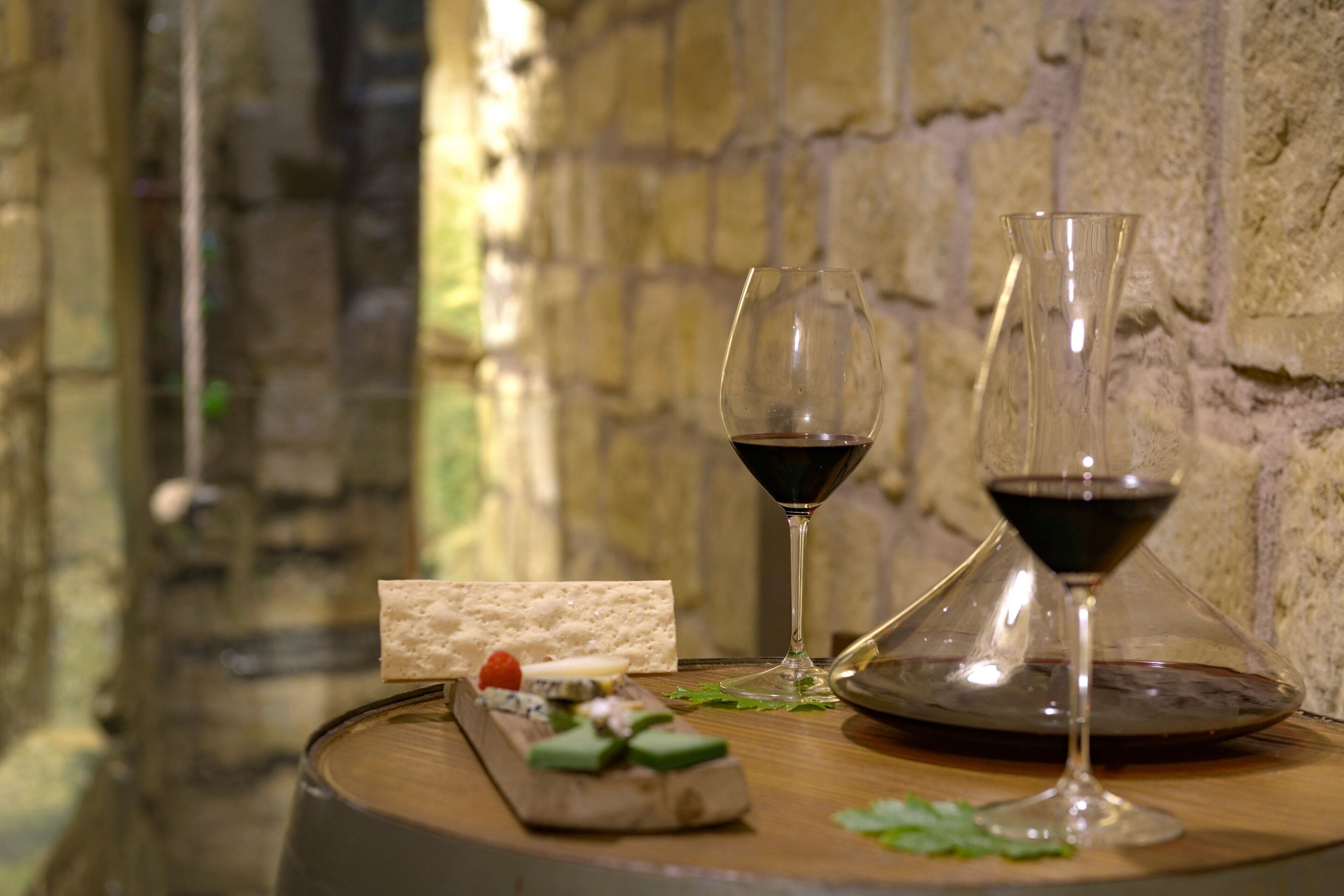 A wooden table with a wine glass, a cheese board with various cheeses and a small piece of bread, and a glass of red wine.