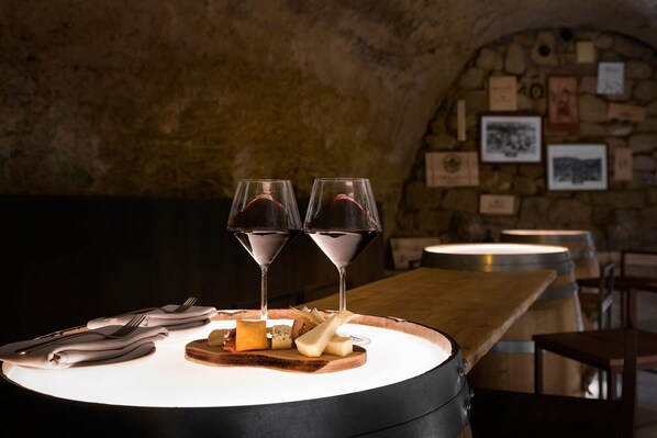 Una mesa con una tabla de madera, queso y copas de vino en una habitación tenuemente iluminada con paredes de piedra y cuadros enmarcados.
