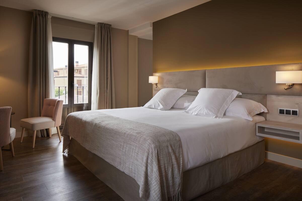 A hotel room with a large bed, two bedside lamps, a chair, and a view of buildings through a window.