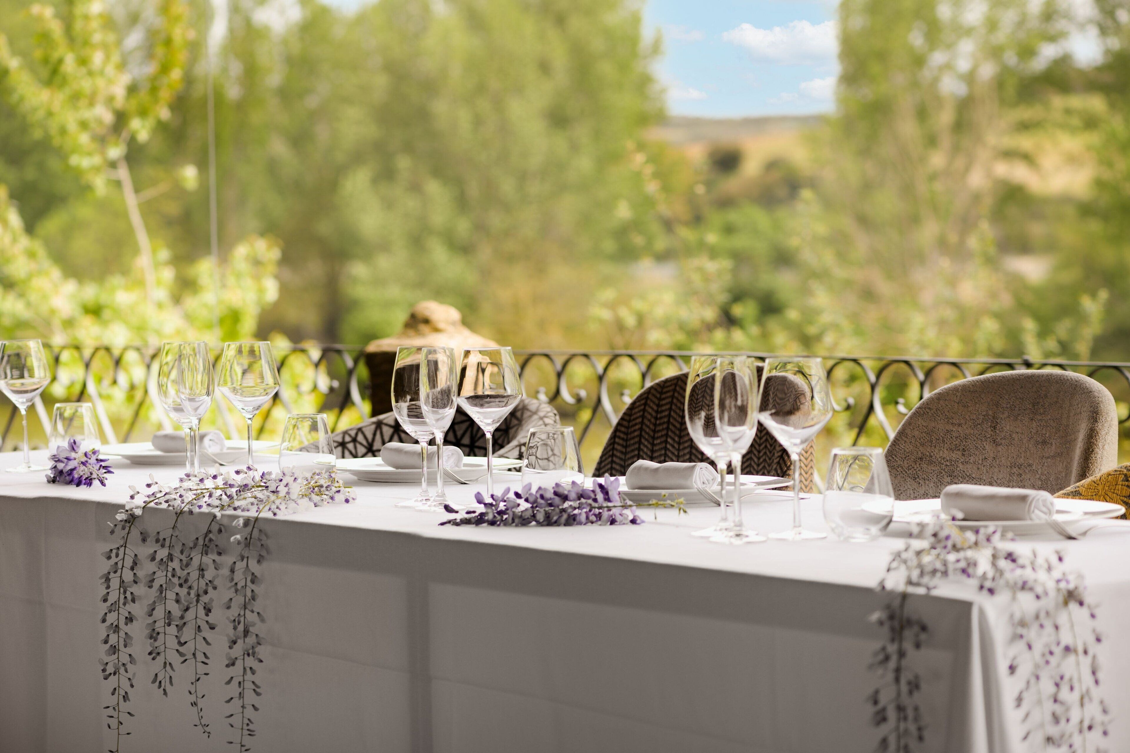 Mesa preparada para una comida con copas de vino, platos y servilleta, con vistas a un área exterior pintoresca con árboles y colinas.