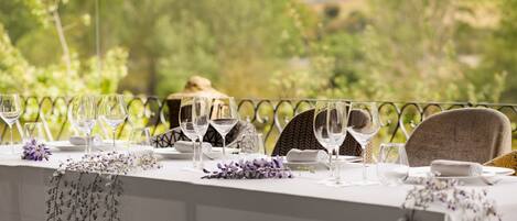 Mesa preparada para una comida con copas de vino, platos y servilleta, con vistas a un área exterior pintoresca con árboles y colinas.