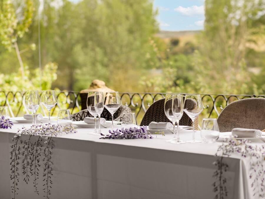 Mesa preparada para una comida con copas de vino, platos y servilleta, con vistas a un área exterior pintoresca con árboles y colinas.