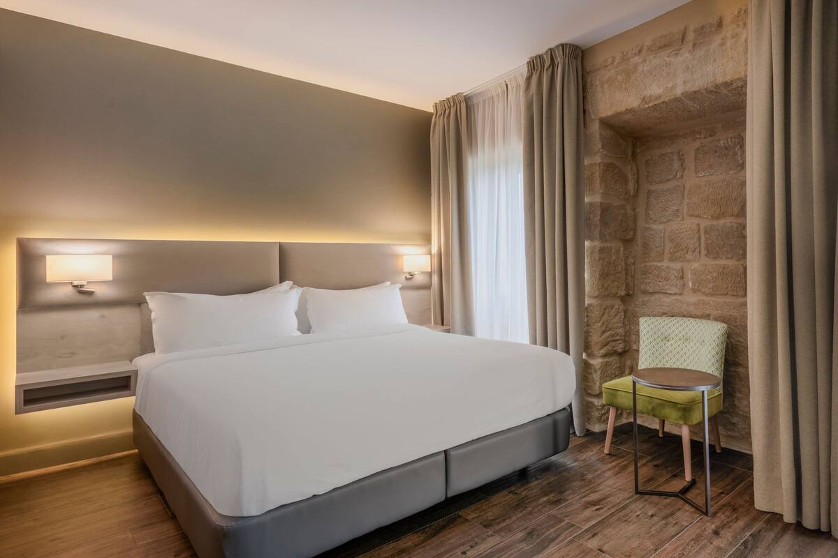 A modern hotel room with a large bed, a nightstand with a lamp, a small green chair, and a stone wall.
