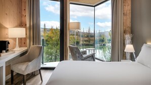 A modern hotel room with a large bed, a desk, and a chair, all facing a scenic view of a lake and mountains. - Palacio Tondon, Autograph Collection (Brinas)