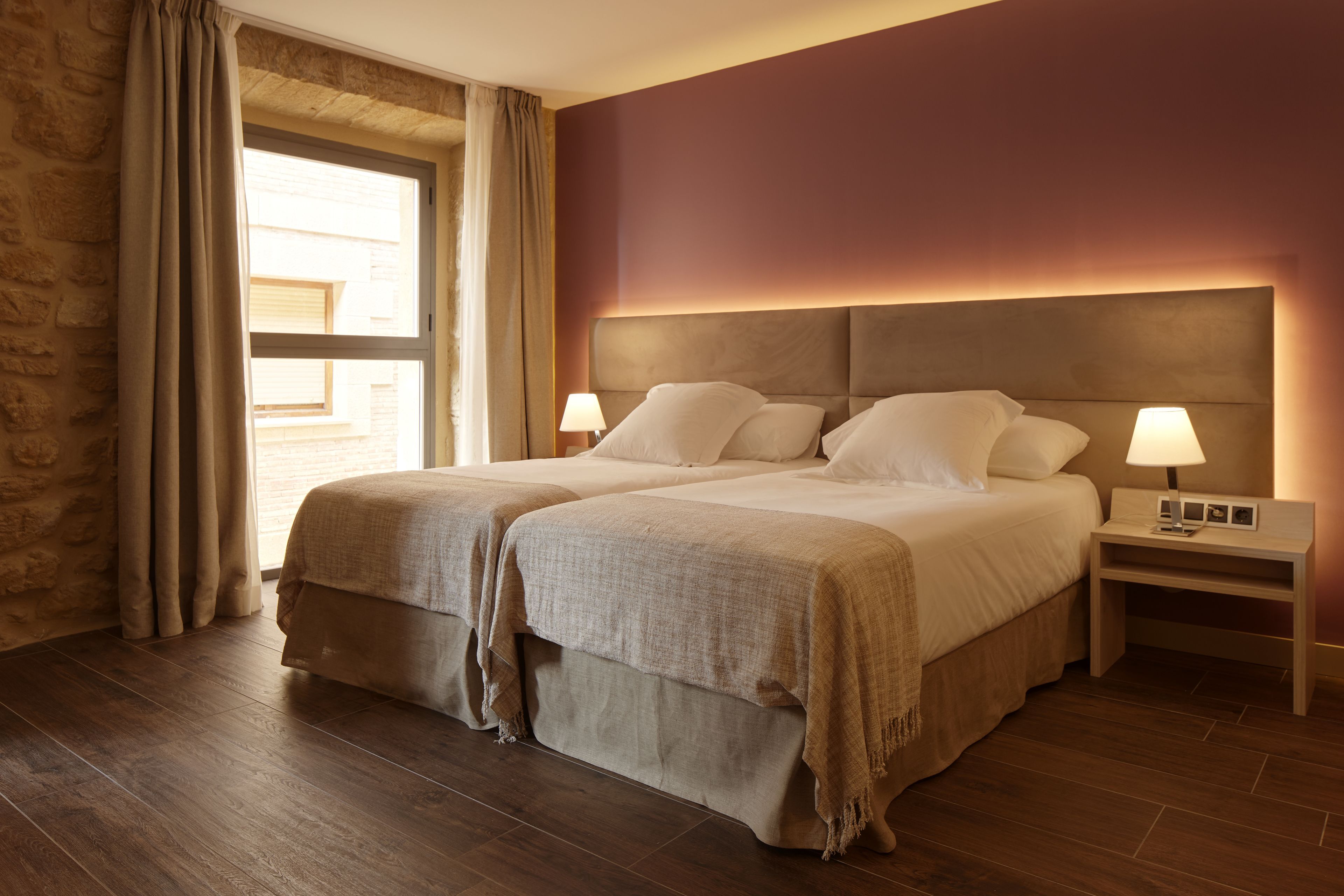 Cama doble con cabecera, dos mesitas de noche con lámparas, una ventana con cortinas y una pared de piedra.