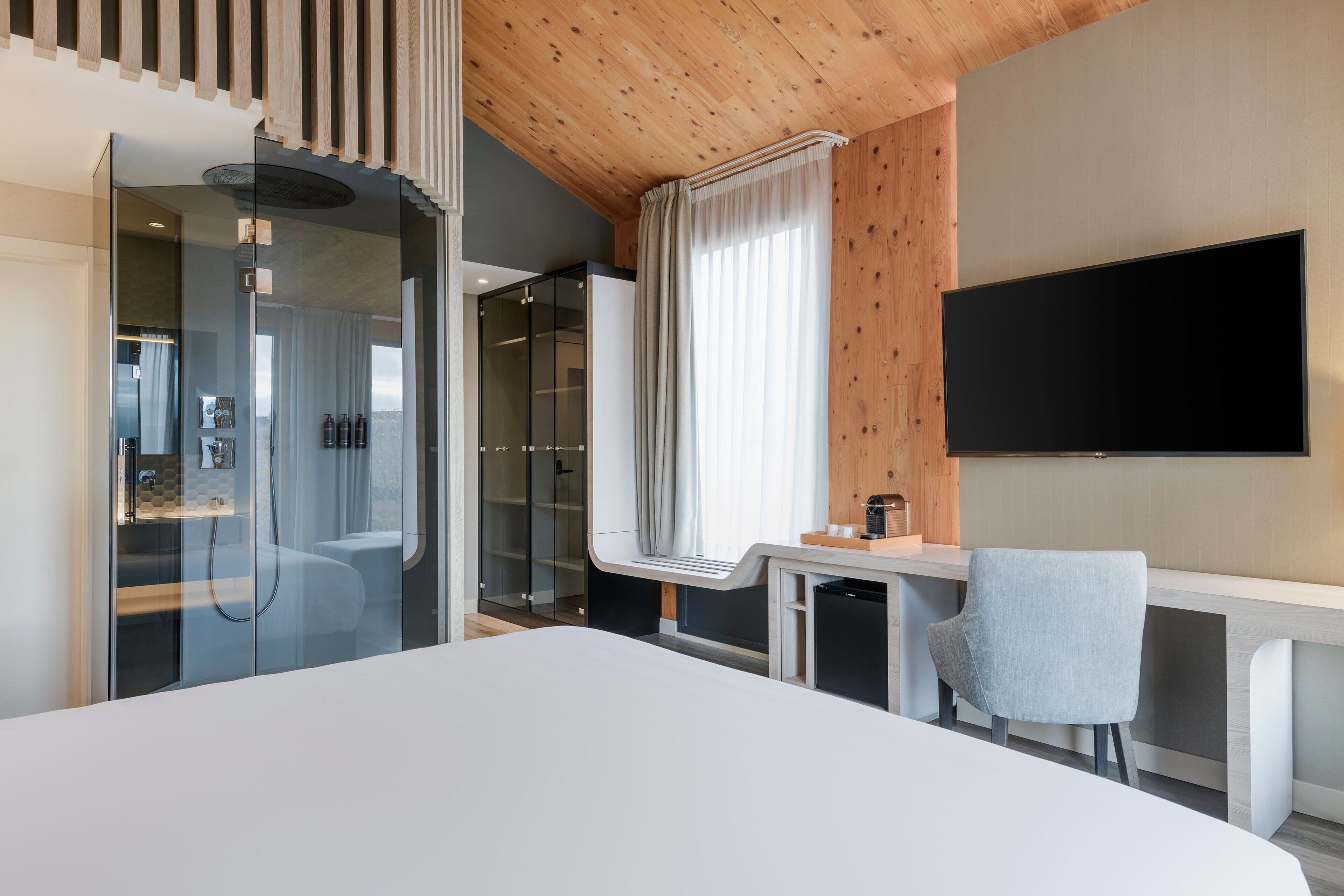 A modern hotel room with a large bed, a desk with a chair, a TV mounted on the wall, and a glass shower enclosure.