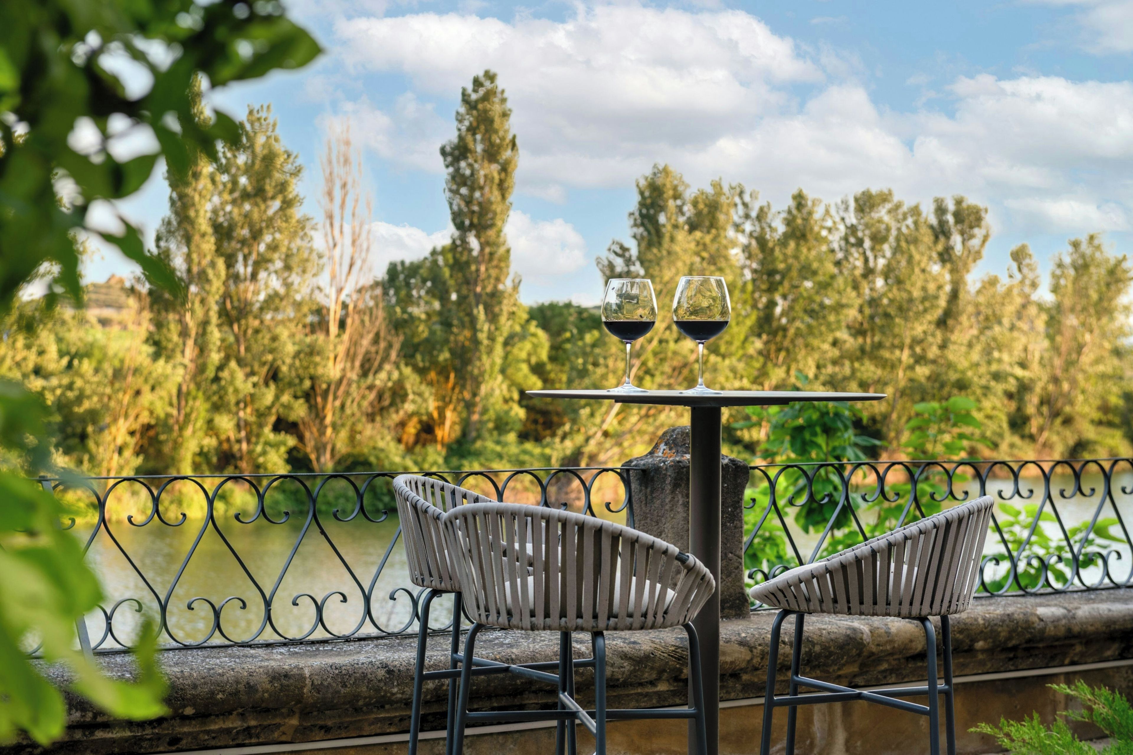 Une table avec deux verres à vin et deux chaises, donnant sur une étendue d’eau, avec des arbres en arrière-plan.