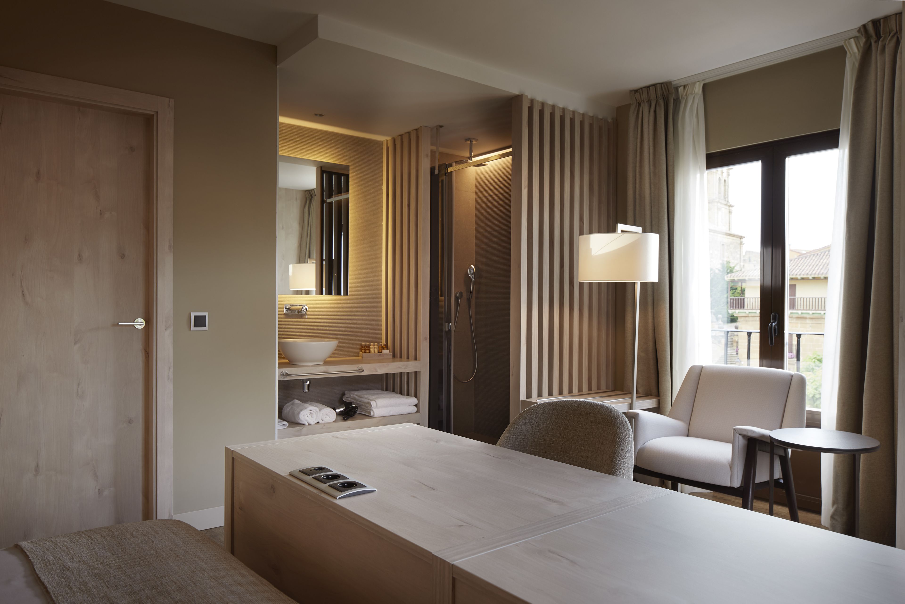 A hotel room with a wooden desk, a chair, a small table, a bathroom with a shower, a sink, and a mirror.