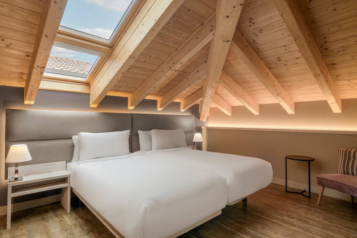 A bedroom with a skylight, a bed, a nightstand, a lamp, and a small table.