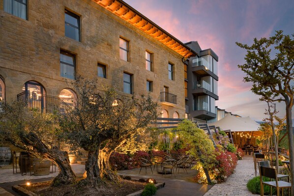 Un patio con un árbol grande, mobiliario para exteriores y un edificio con varias ventanas y balcones.