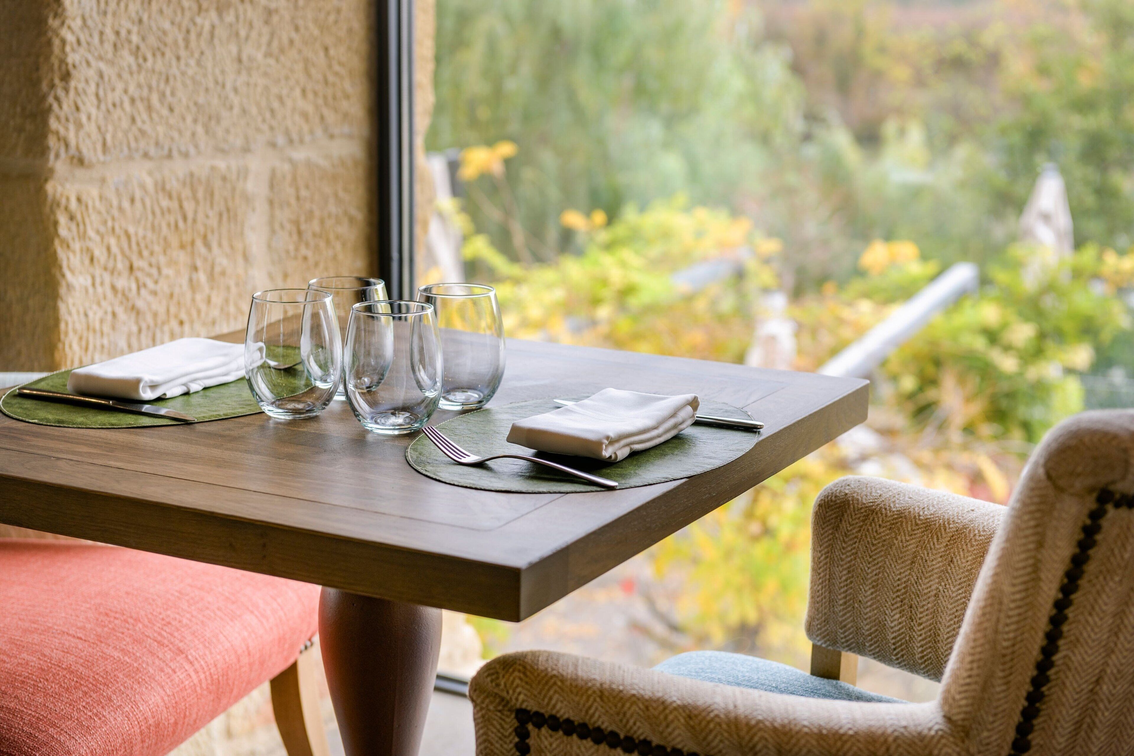 Mesa de comedor dispuesta con vasos transparentes, un servilleta doblada y cubiertos, con vistas a la vegetación exterior.