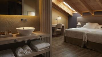 A modern bathroom with a large sink, mirror, and towel rack. Adjacent is a bedroom with a desk, chair, and two beds.