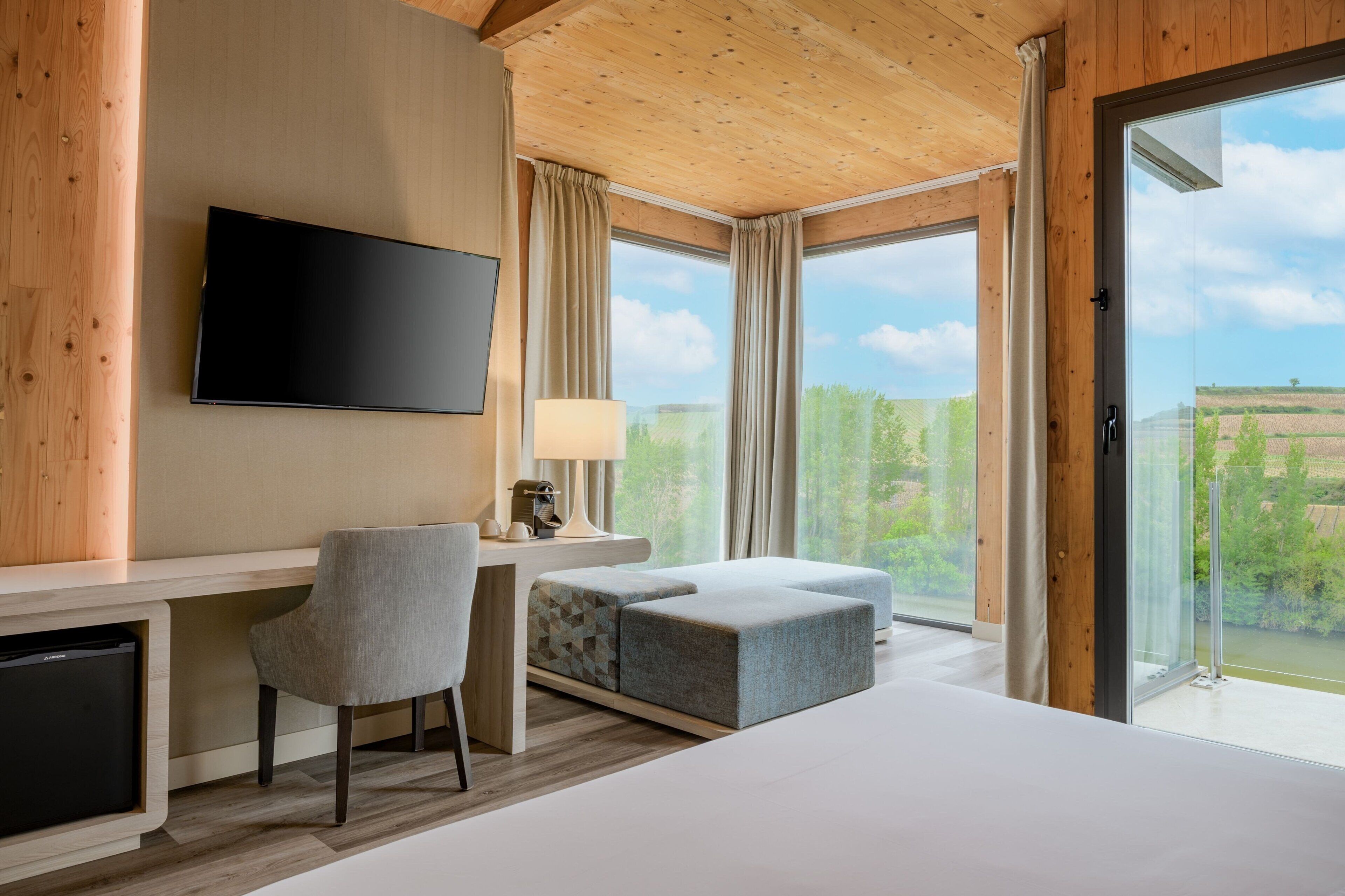 Una habitación de hotel moderna con una cama grande, un escritorio con silla, televisión y vistas al exterior a través de amplias ventanas.