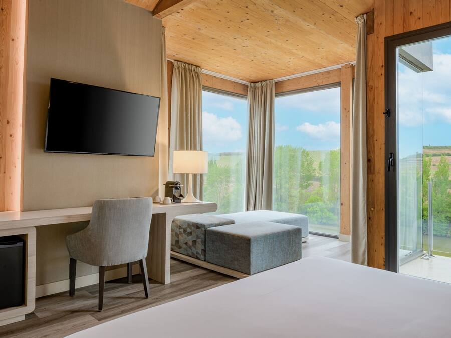 Una habitación de hotel moderna con una cama grande, un escritorio con silla, televisión y vistas al exterior a través de amplias ventanas.