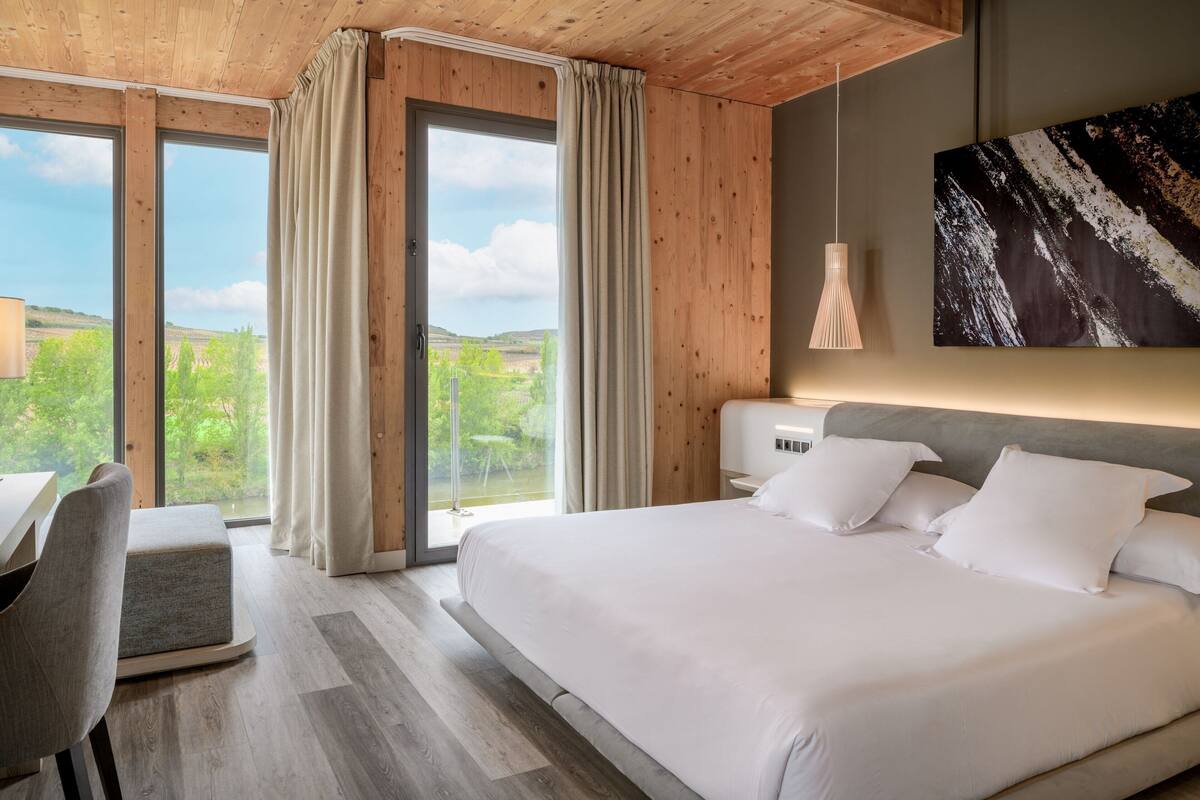 A modern bedroom with a large bed, a desk, and a view of the outdoors.