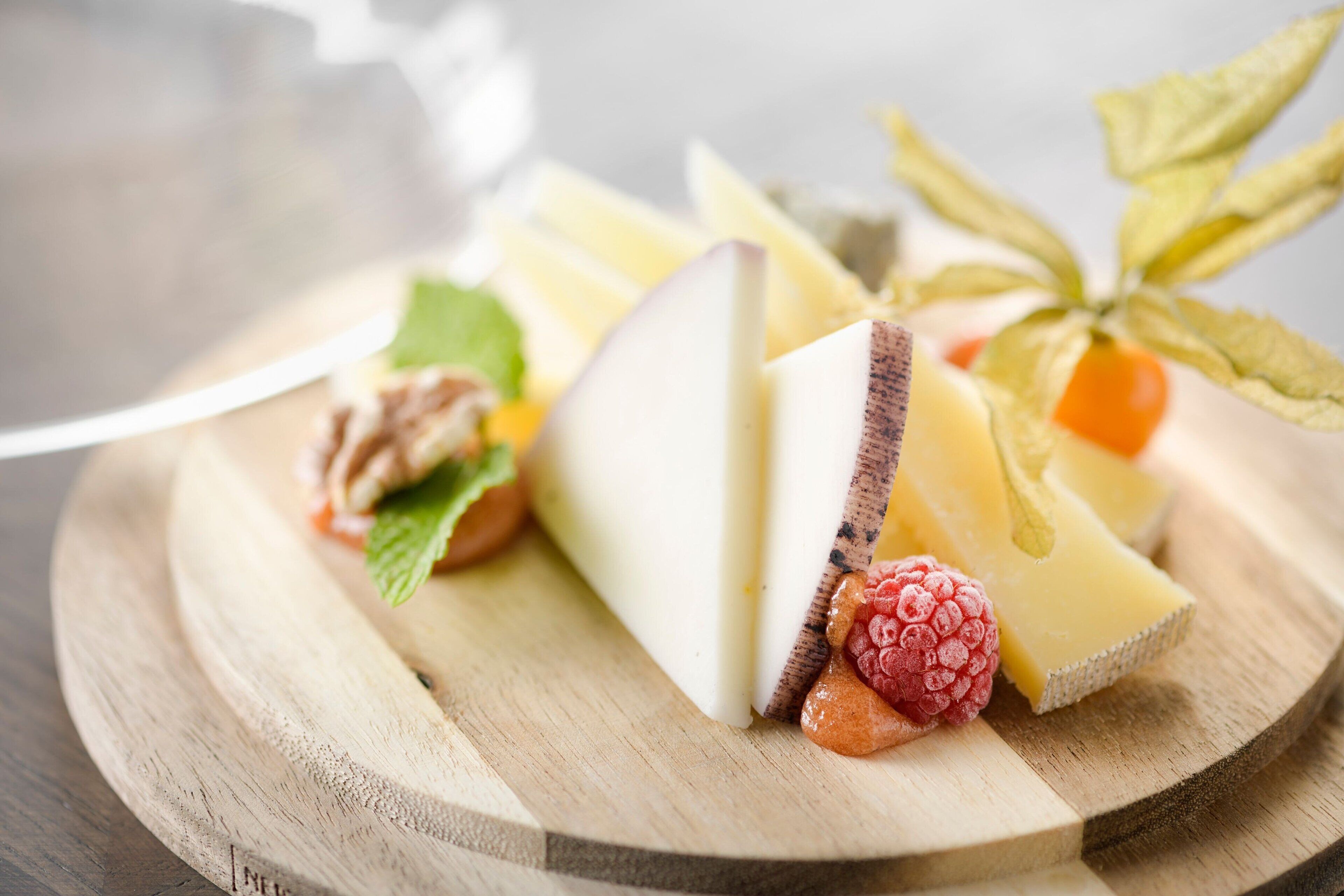 A wooden board with various cheeses, a raspberry, and a leaf.