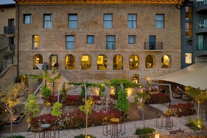 Un edificio de piedra con amplias ventanas de arco y un patio con mobiliario exterior y plantas.