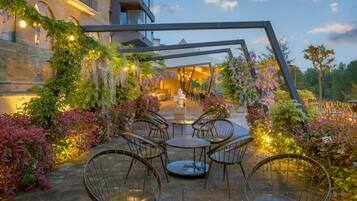 A courtyard with outdoor seating, surrounded by lush greenery and flowering plants, under a clear sky.