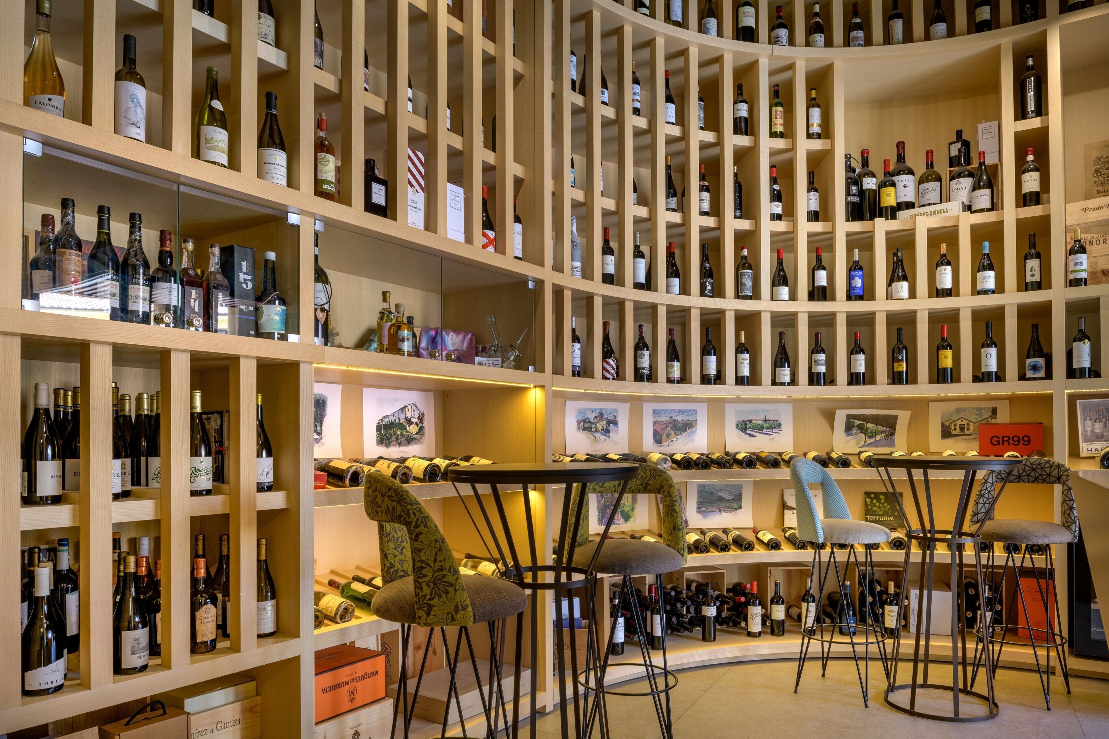 Una bodega con múltiples estantes de botellas de vino, una zona de bar con taburetes y cuadros enmarcados en las paredes.