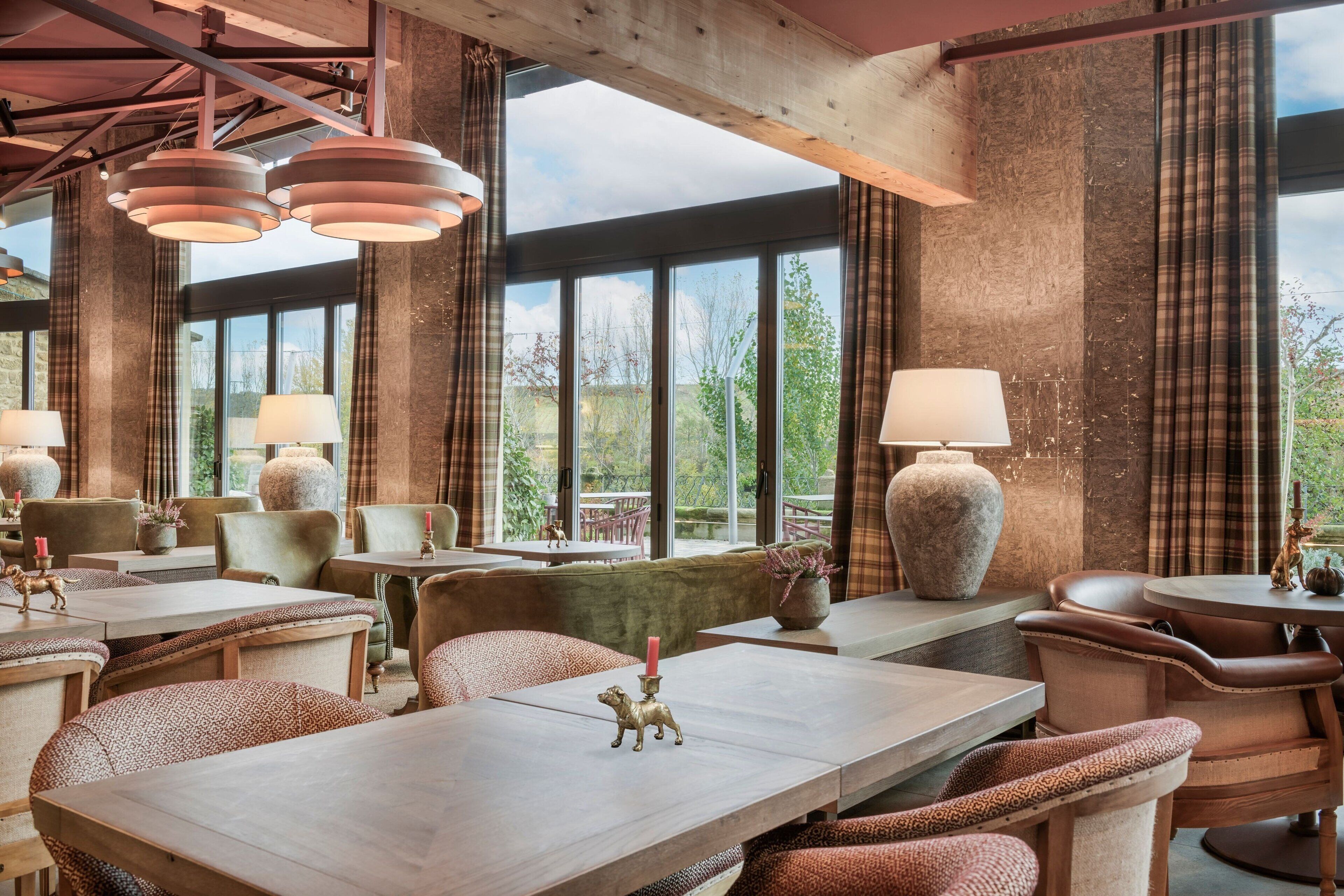 A restaurant with high ceilings, large windows, and a rustic interior. Tables are arranged with cushioned chairs, and there are decorative vases and candles on the tables.