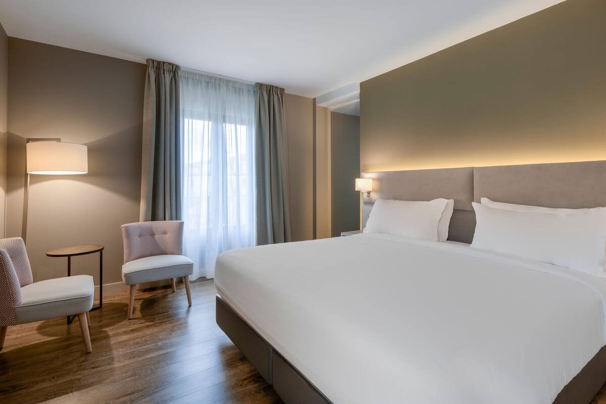 A hotel room with a large bed, two chairs, a small table, and a lamp.