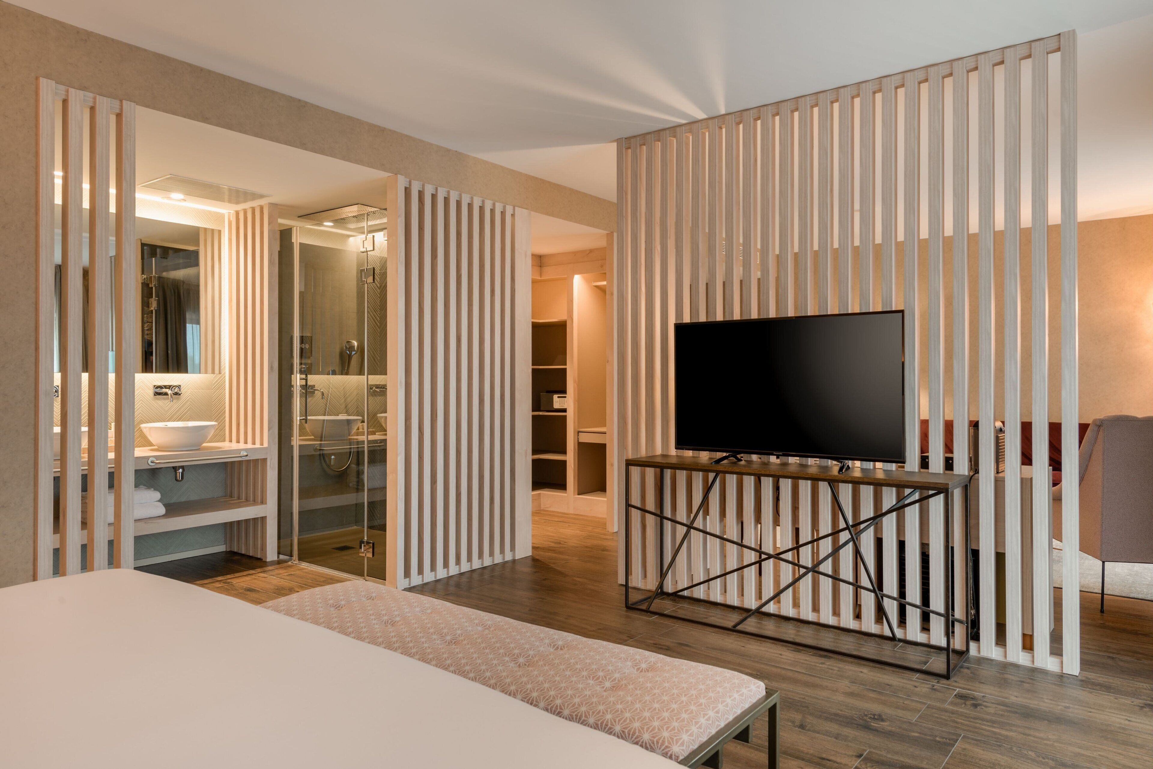 A modern hotel room with a large bed, a TV on a stand, a bathroom with a glass shower enclosure, and a wooden partition.