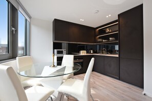 A modern kitchen with dark cabinetry, a glass dining table, and white chairs. - Hoxton by Servprop (London)