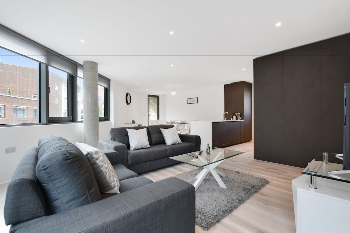 A modern living room with a grey sofa, a glass coffee table, and a large window offering a view of a brick building.