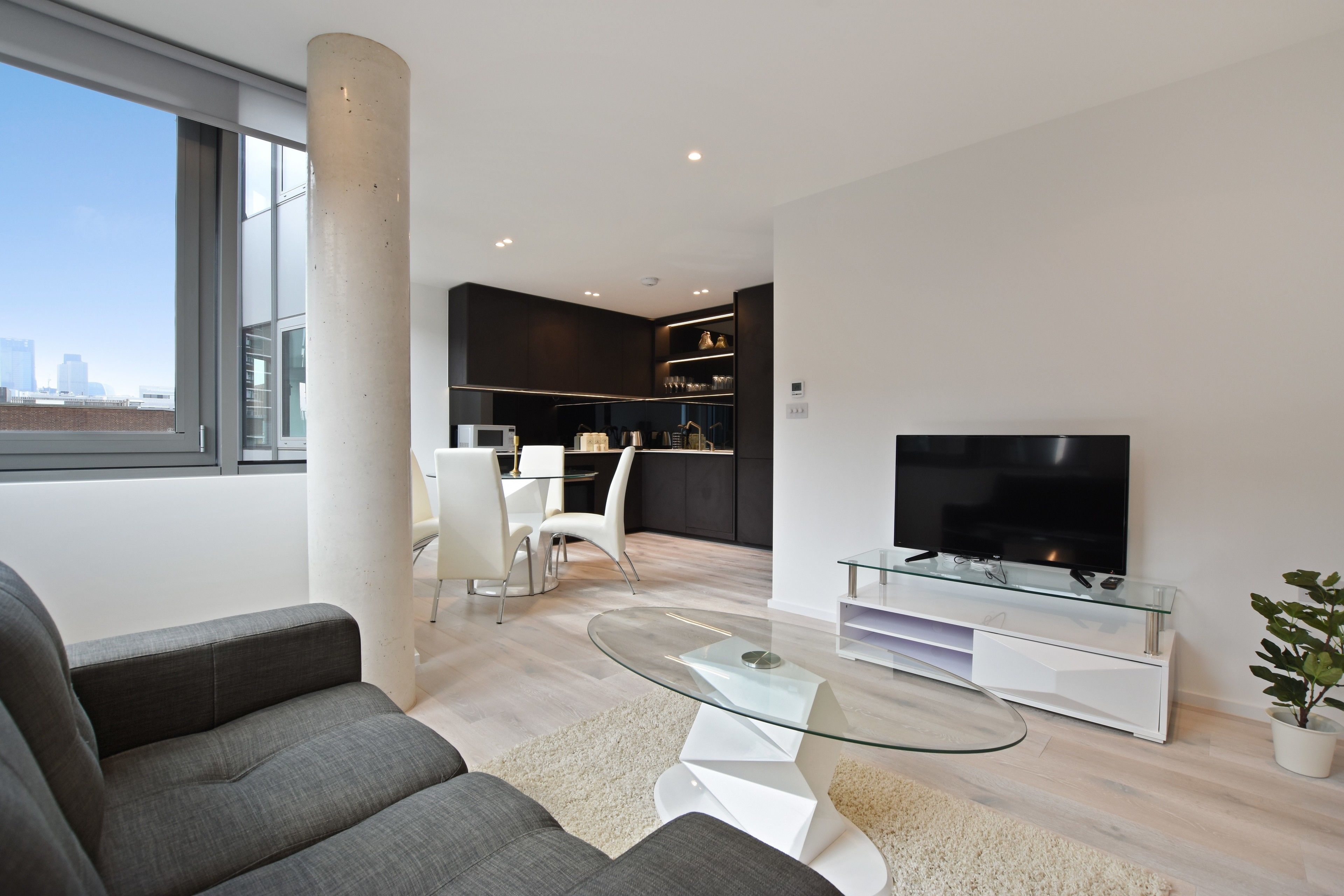 A modern living room with a sofa, a glass coffee table, and a TV on a glass stand. There is a dining area with a white table and chairs, and a kitchen in the background.