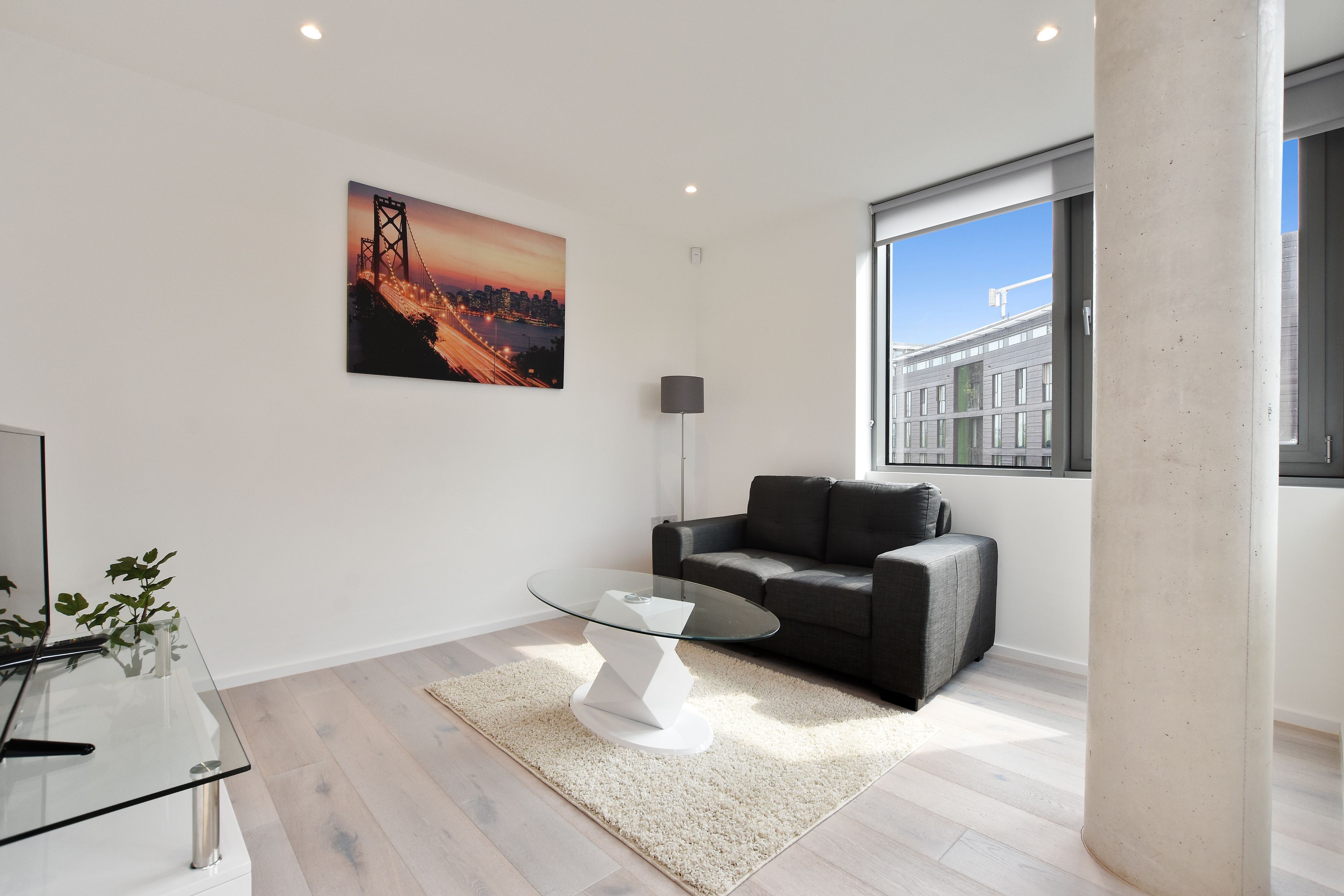 A modern living room with a glass coffee table, a grey sofa, a cityscape painting, and a large window offering a view of buildings.