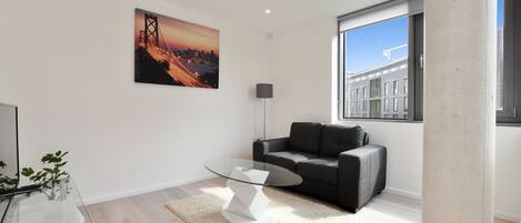 A modern living room with a glass coffee table, a grey sofa, a cityscape painting, and a large window offering a view of buildings.