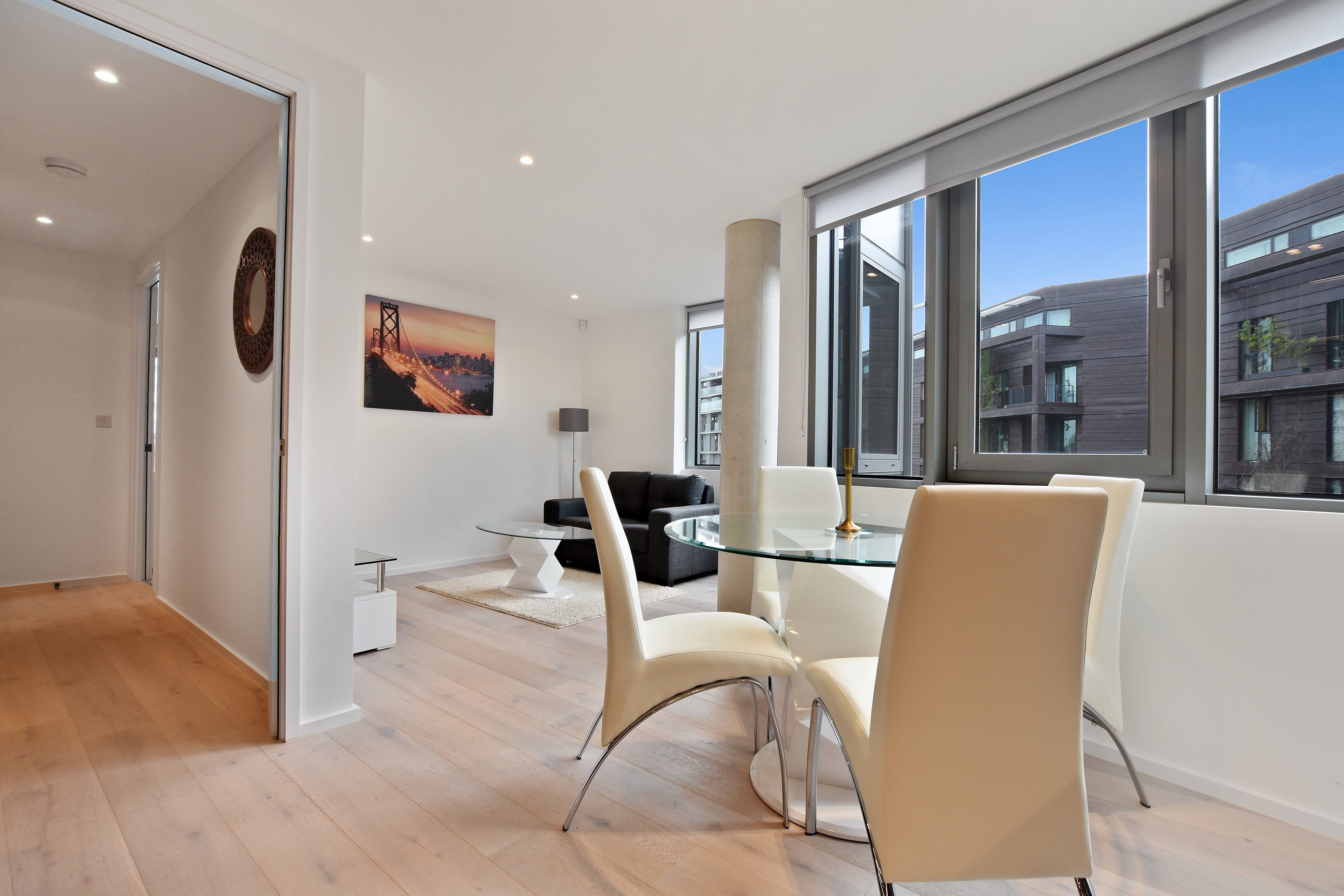 Et moderne spiseområde med et glasbord og hvide stole, en sort sofa og et bybillede på væggen.