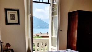 A bedroom with a large bed, a view of a mountain and lake through an open window, a framed picture on the wall, and a wooden wardrobe. - Villa Laura (Lierna)