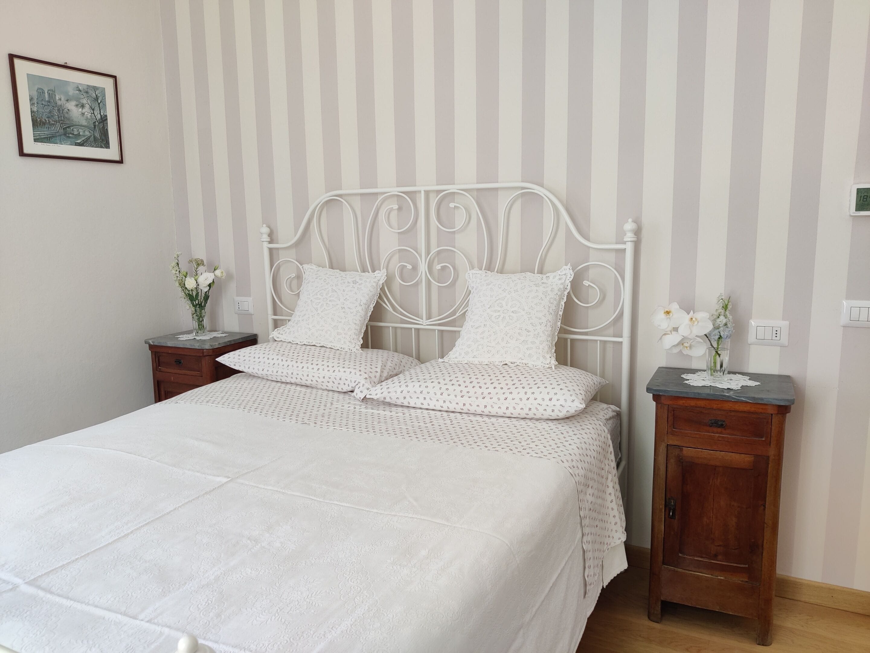 A neatly made bed with a white bedspread and pillows, a metal headboard, and two bedside tables with floral vases.