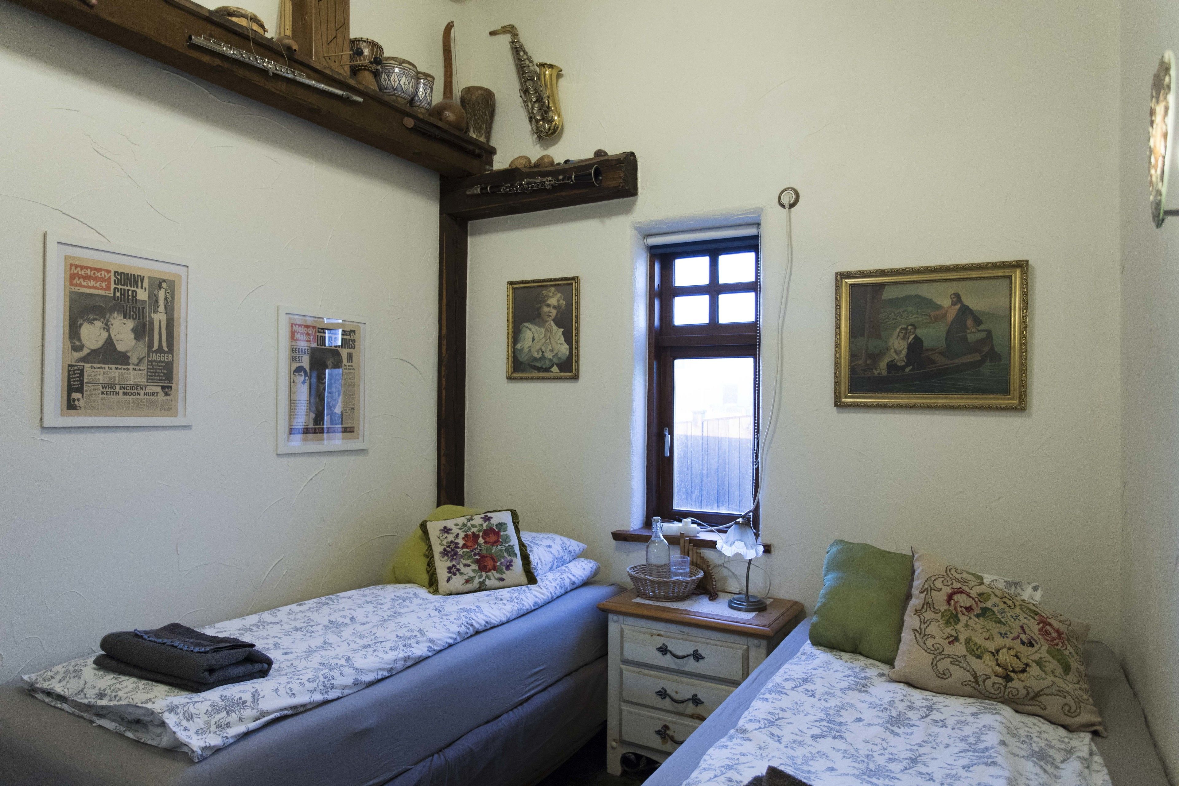 A small, cozy bedroom with two single beds, a nightstand, a window, and various wall decorations including framed pictures and a poster.