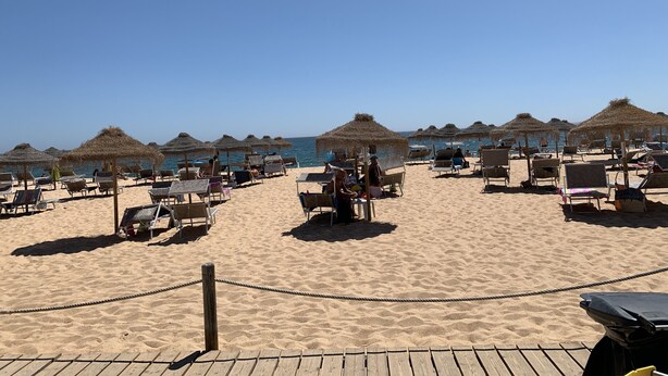 Une plage bordée de rangées de chaises longues et d’ombrelles en chaume, une terrasse en bois au premier plan et la mer en arrière-plan.