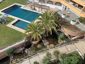 Une piscine avec des chaises longues, une pergola, des palmiers et un petit pont enjambant un bassin d’eau décoratif.