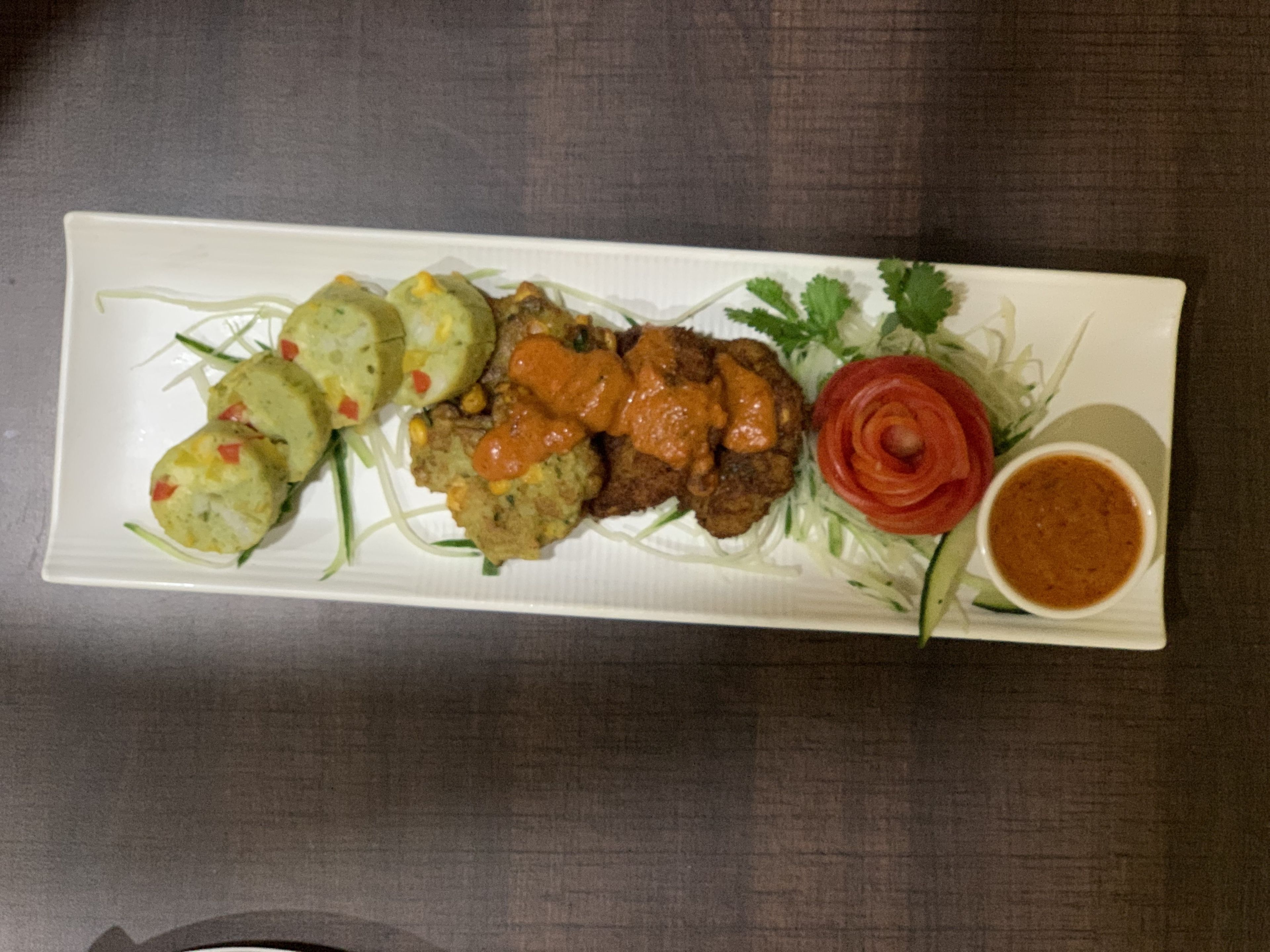 A rectangular white plate with various food items including patties, a slice of tomato, and a small bowl of sauce.
