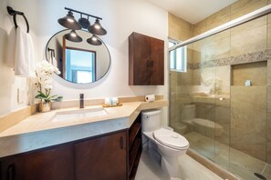 A modern bathroom with a marble countertop, a round mirror, a wooden cabinet, a toilet, and a glass-enclosed shower.