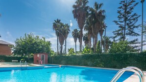 A clear blue swimming pool with metal ladders, surrounded by palm trees and a hedge. - HomeHolidaysRentals Apartamento Amaral - Costa Barcelona (Pineda de Mar)