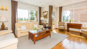 A spacious living room with a sofa, armchairs, a coffee table, and a desk. Large windows provide natural light and views of greenery outside. - HomeHolidaysRentals Apartamento Amaral - Costa Barcelona (Pineda de Mar)