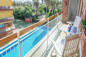 A balcony with a pool, white chairs, and a view of a residential area with palm trees and a red building. - HomeHolidaysRentals Apartamento Amaral - Costa Barcelona (Pineda de Mar)