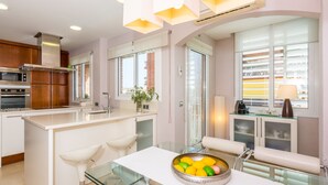 A modern kitchen with a central island, white cabinetry, and a glass dining table with a bowl of fruit. - HomeHolidaysRentals Apartamento Amaral - Costa Barcelona (Pineda de Mar)