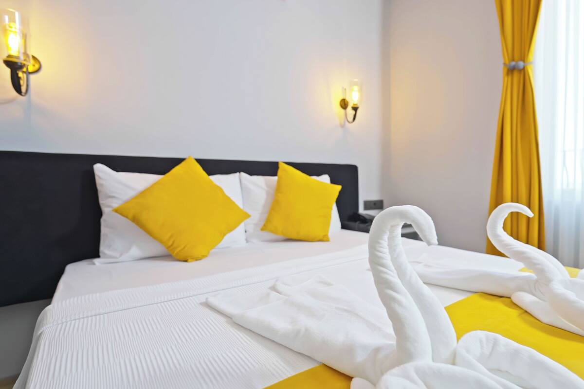 A hotel room with a bed, two yellow pillows, a white headboard, and a yellow curtain.