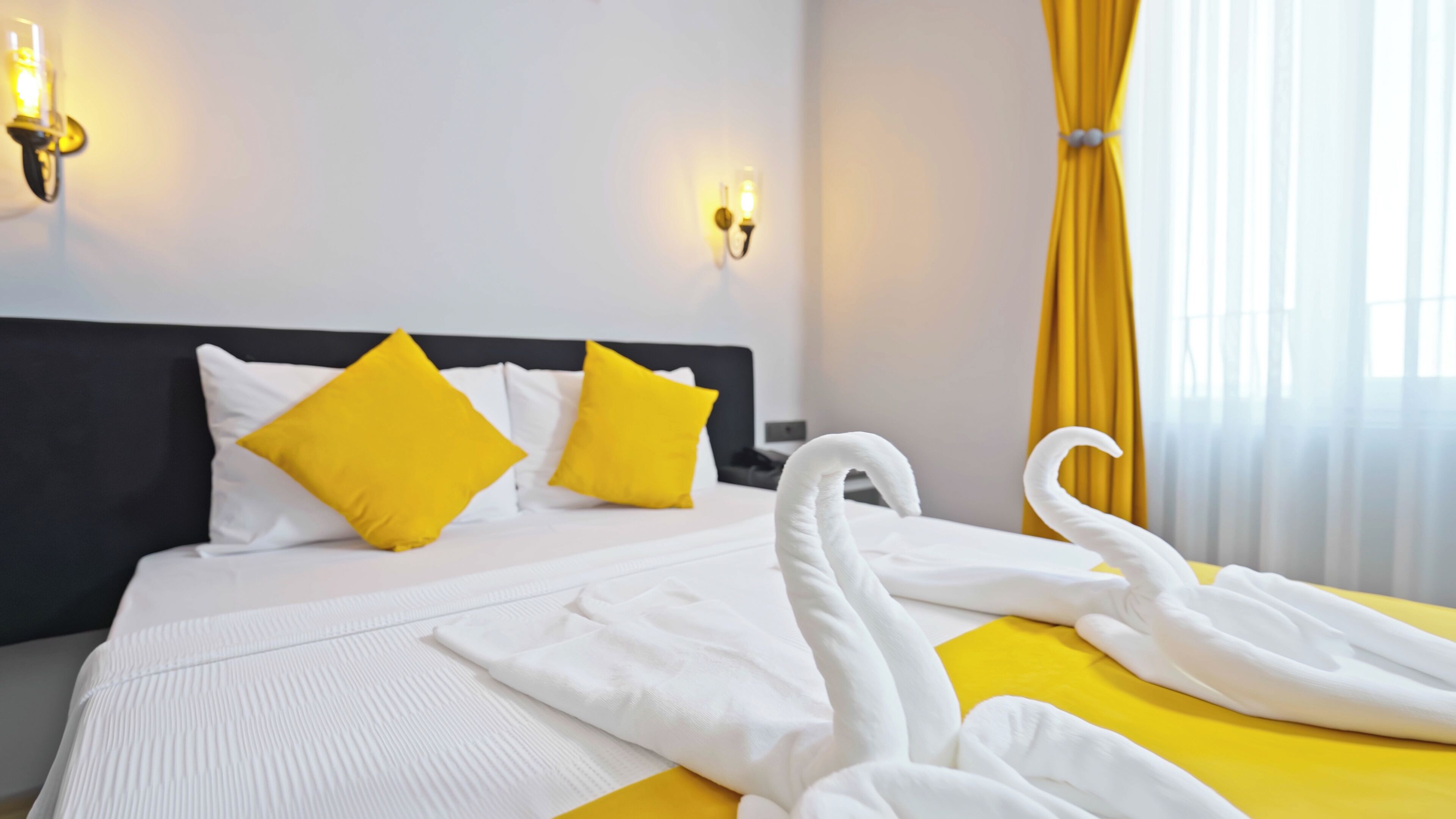 A hotel room with a bed, two yellow pillows, a white headboard, and a yellow curtain.