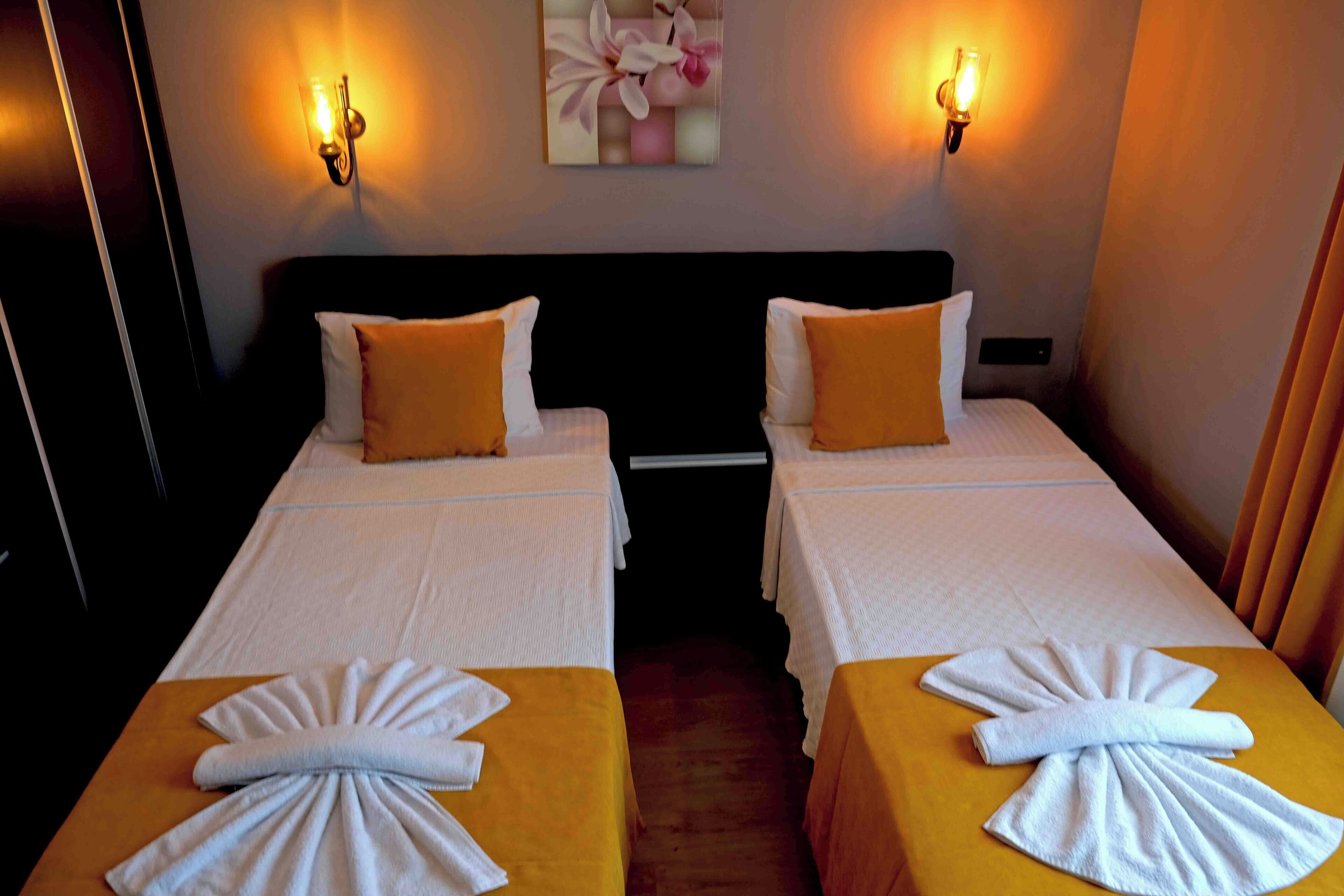 A hotel room with two beds, each with a white bedspread and a yellow accent pillow. There are wall-mounted lights and a painting above the beds.