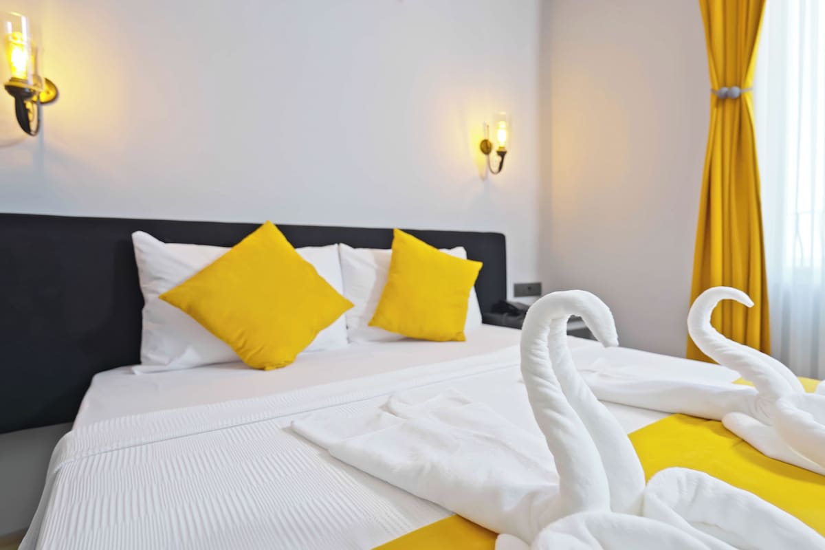 Une chambre d’hôtel avec un lit, deux oreillers jaunes, une tête de lit blanche et un rideau jaune.