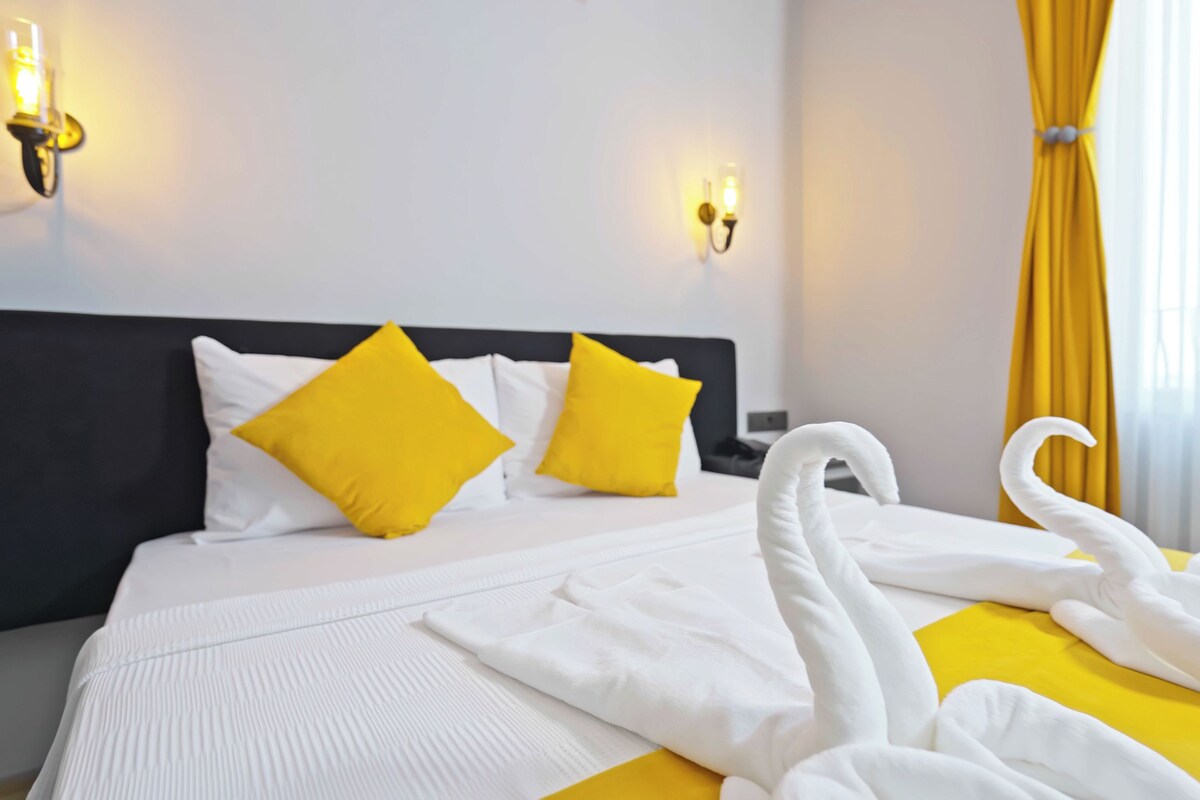 A hotel room with a bed, two yellow pillows, a white headboard, and a yellow curtain.