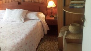 Une pièce chaleureuse avec un lit en bois, une lampe de chevet, une petite étagère avec des livres et un chapeau, et un tapis à motifs.