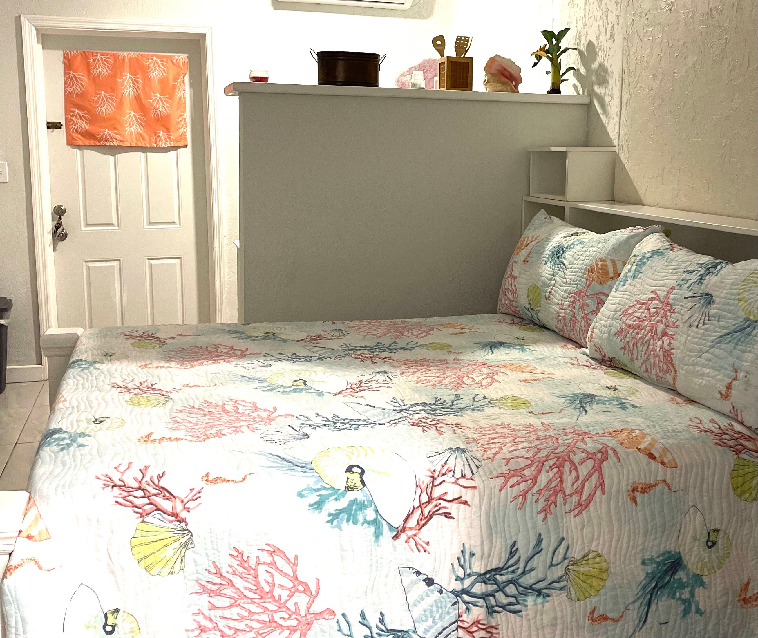 A bedroom with a bed covered in a floral-patterned quilt, a white door, and a shelf with decorative items.
